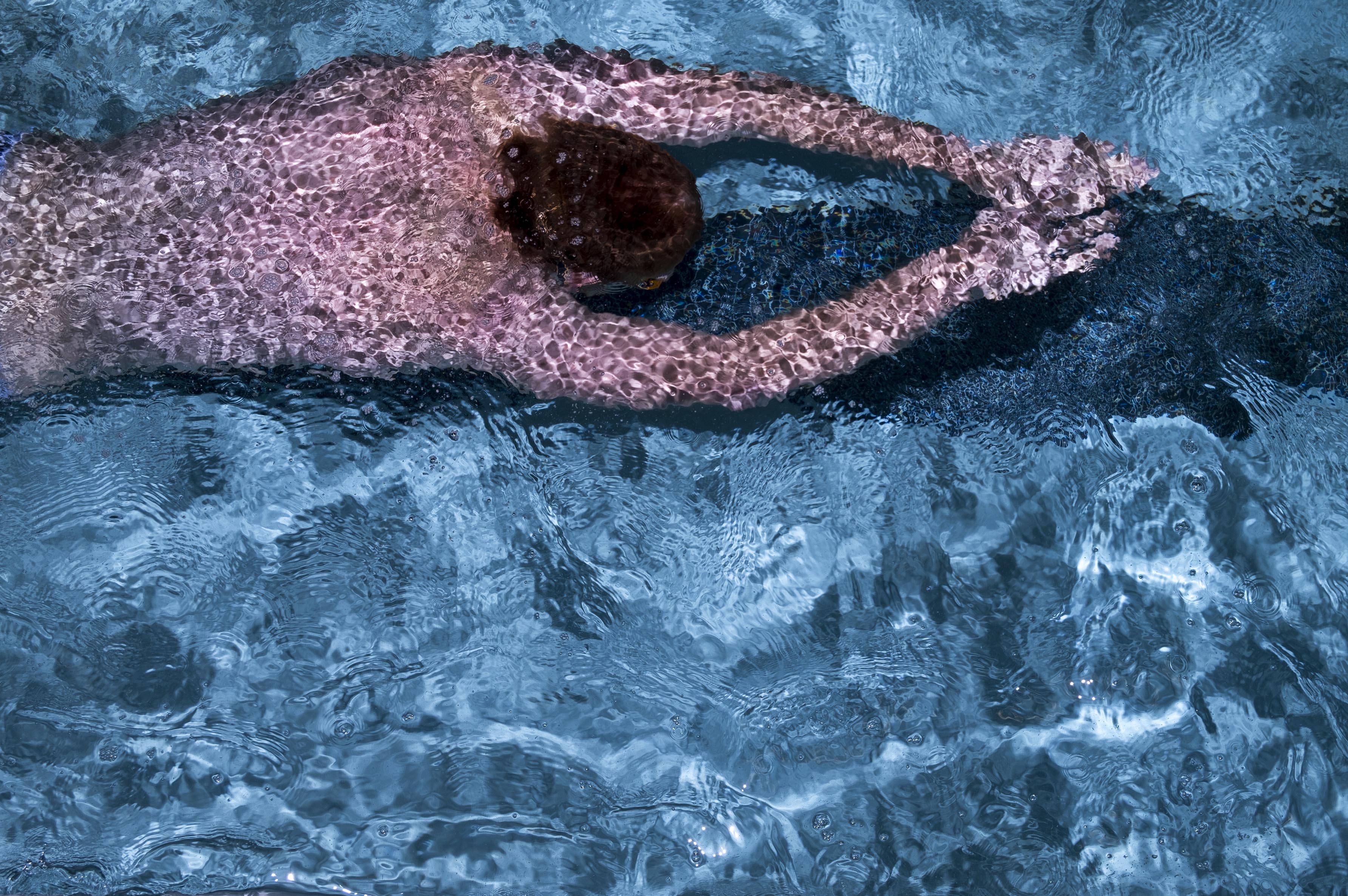 A photograph of a man in a speedo swimming in a pool with his arms in front of him, shot from above.