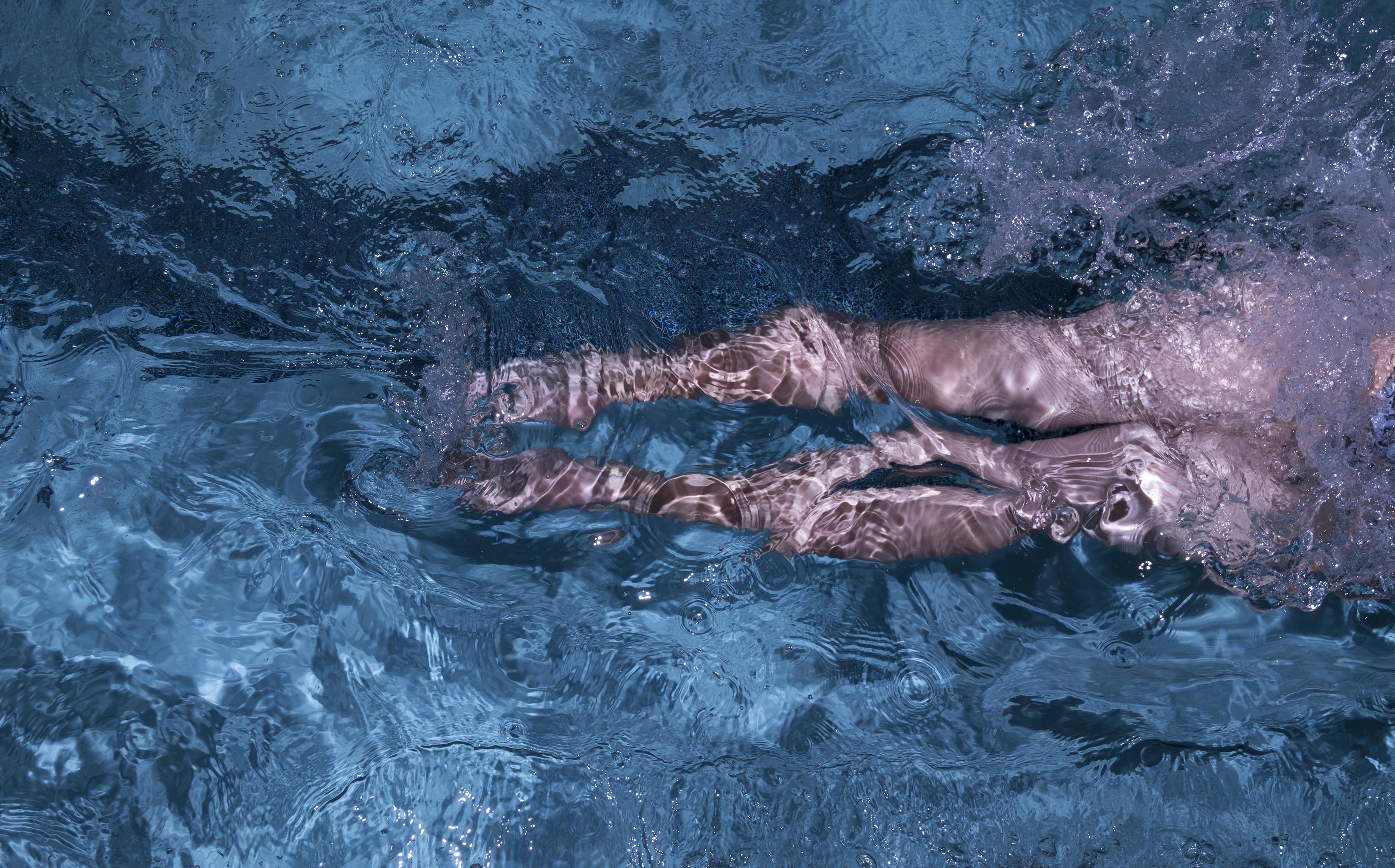 A photograph of a woman's legs disappearing to the right as she swims in a pool lane, splashing lots of water, shot from above.