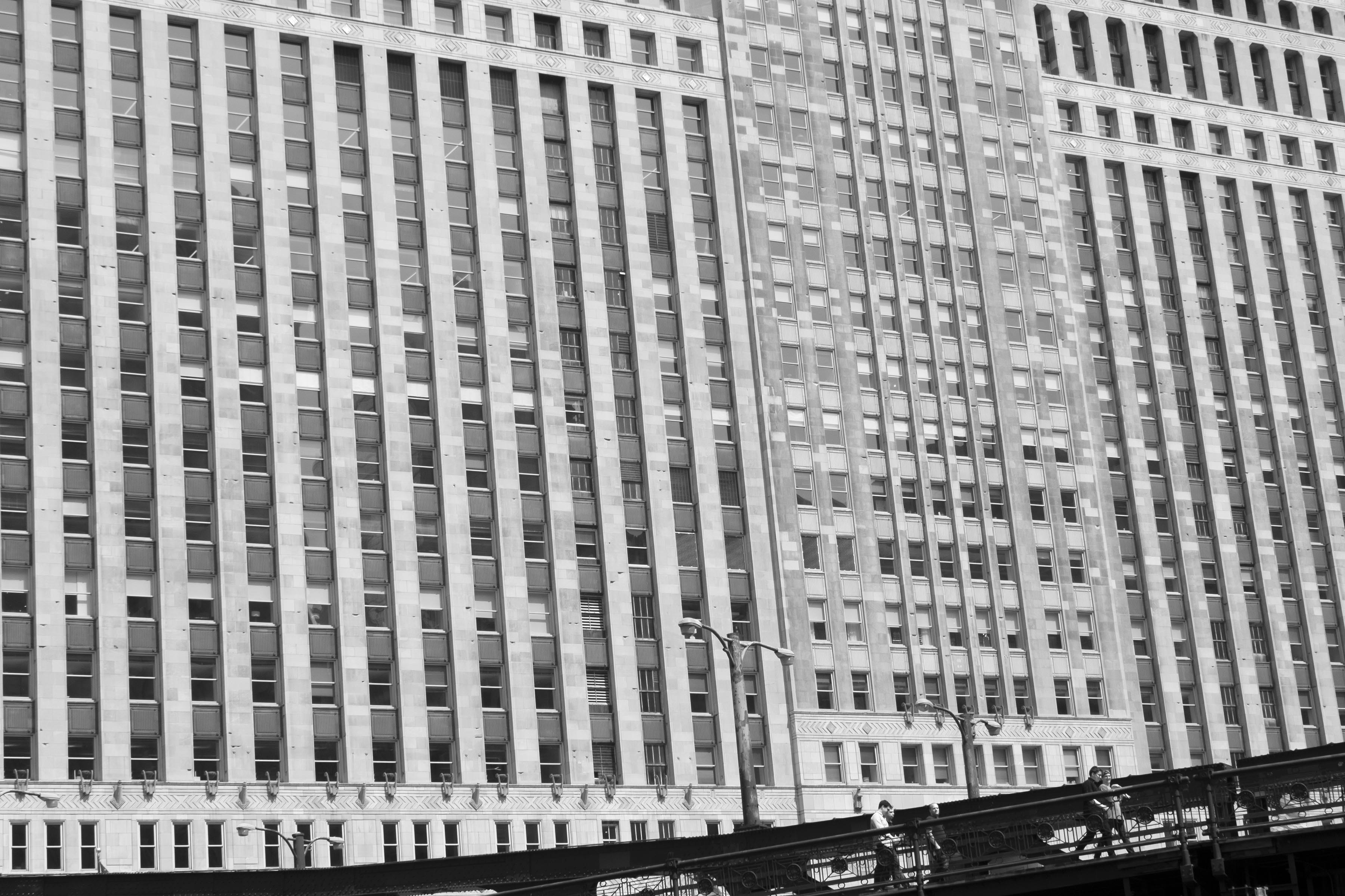 A photograph of a massive building facade with a grid of windows. The right bottom corner of the photograph features a sliver of dark bridge, with 3 small human figures walking on it.